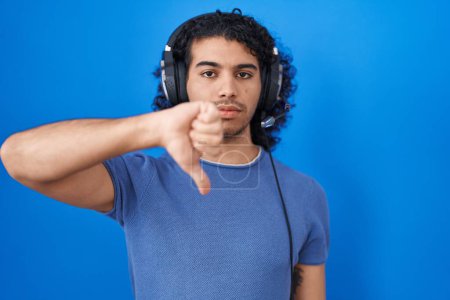 Photo for Hispanic man with curly hair listening to music using headphones looking unhappy and angry showing rejection and negative with thumbs down gesture. bad expression. - Royalty Free Image