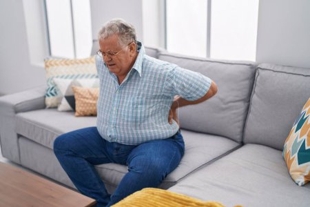 Téléchargez les photos : Homme aux cheveux gris d'âge moyen souffrant de maux de dos assis sur le canapé à la maison - en image libre de droit