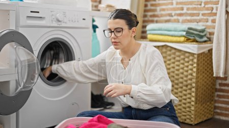 Foto de Joven mujer hispana hermosa poniendo ropa en la lavadora en la sala de lavandería - Imagen libre de derechos