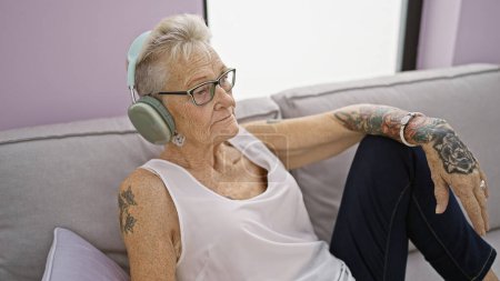 Téléchargez les photos : Femme âgée aux cheveux gris écoutant de la musique assise sur un canapé à la maison - en image libre de droit