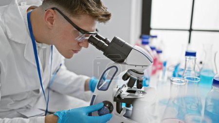 Photo for Handsome young caucasian man, a dedicated scientist, deeply engrossed in analyzing samples under the microscope at a professional medical lab. - Royalty Free Image