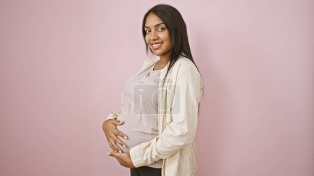 Foto de Joven embarazada alegre con una sonrisa radiante disfruta tocando alegremente su vientre, de pie casualmente sobre un fondo rosa aislado - Imagen libre de derechos