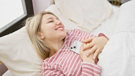 Photo for Smiling young woman relaxing in bedroom holding smartphone, portraying leisure and technology at home. - Royalty Free Image
