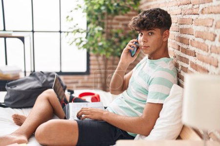 Téléchargez les photos : Jeune adolescent hispanique parlant sur smartphone en utilisant un ordinateur portable assis sur le lit dans la chambre - en image libre de droit