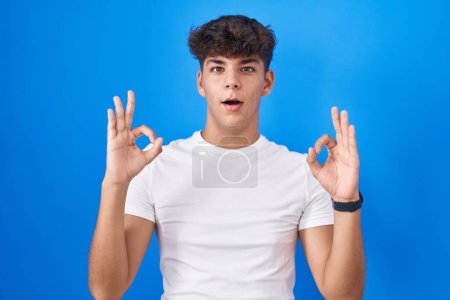 Téléchargez les photos : Adolescent hispanique debout sur fond bleu regardant surpris et choqué faire ok symbole d'approbation avec les doigts. expression folle - en image libre de droit