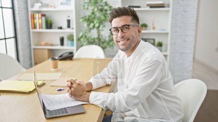 Photo for Handsome young hispanic man, confidently reading business documents with a successful smile, working at his office desk indoors. - Royalty Free Image