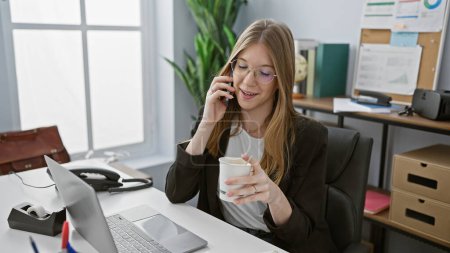 Téléchargez les photos : Femme blonde parlant au téléphone et tenant la tasse dans un bureau moderne avec un ordinateur portable et des accessoires de bureau - en image libre de droit
