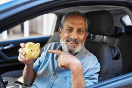 Foto de Hombre mayor sosteniendo alcancía sonriendo feliz señalando con la mano y el dedo en el coche - Imagen libre de derechos