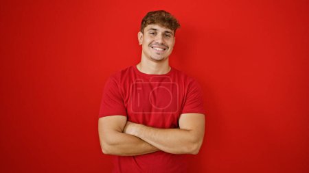 Téléchargez les photos : Jeune homme hispanique joyeux qui respire la confiance, debout avec les bras croisés, rayonnant un sourire sur un mur rouge isolé, rayonnant de positivité, de joie et de rire contagieux - en image libre de droit