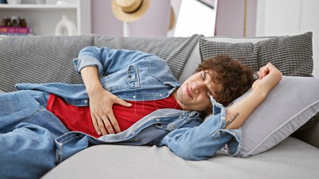 Photo for Hispanic man with curly hair, wearing denim shirt, feeling stomach pain on a grey sofa at home. - Royalty Free Image
