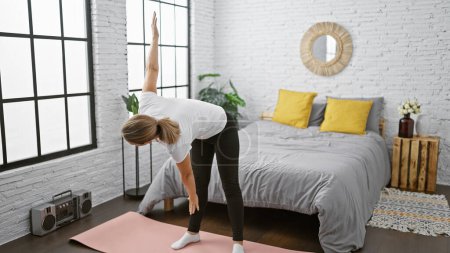 Photo for Serious young blonde woman finding balance, strengthening her concentration with morning yoga exercise on her bed, warming up in cozy indoor bedroom space, enjoying calmness and relaxation. - Royalty Free Image
