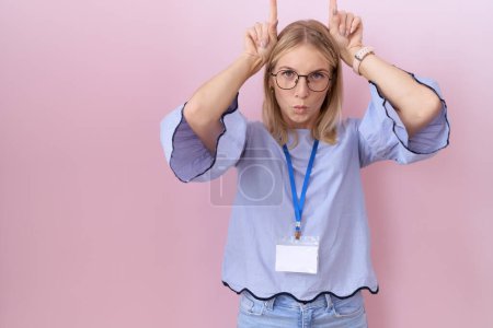 Téléchargez les photos : Jeune femme d'affaires caucasienne portant une carte d'identité faisant un geste drôle avec le doigt au-dessus de la tête comme des cornes de taureau - en image libre de droit