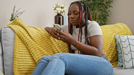Téléchargez les photos : Belle femme afro-américaine, détendue à la maison, se concentre intensément sur la dactylographie d'un message sur son téléphone alors qu'elle est assise sur le canapé. - en image libre de droit