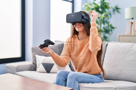 Foto de Joven mujer hispana hermosa jugando videojuego usando gafas de realidad virtual y joystick en casa - Imagen libre de derechos