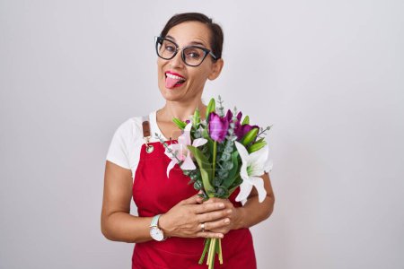 Photo for Middle age brunette woman wearing apron working at florist shop holding bouquet sticking tongue out happy with funny expression. emotion concept. - Royalty Free Image