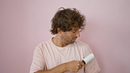 Foto de Un hombre joven casual ellos con el pelo rizado sosteniendo un rodillo de pelusa sobre un fondo aislado rosa. - Imagen libre de derechos