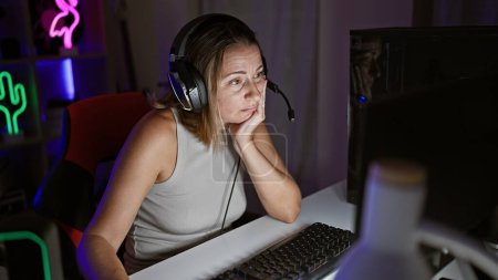 Téléchargez les photos : Épuisé jeune femme streamer blonde sentir la tension du jeu sans fin, assis dans la salle de jeux sombre la nuit, immergé dans la réalité virtuelle via ordinateur et casque - en image libre de droit