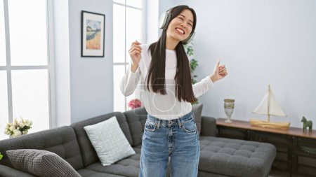 Foto de Una mujer asiática alegre que usa auriculares baila sola en una sala de estar moderna, expresando felicidad sin preocupaciones y entretenimiento en el hogar. - Imagen libre de derechos