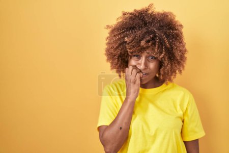 Photo for Young hispanic woman with curly hair standing over yellow background looking stressed and nervous with hands on mouth biting nails. anxiety problem. - Royalty Free Image