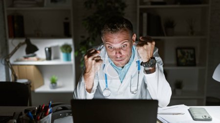 Photo for Stressed doctor in clinic office working late with computer and paperwork - Royalty Free Image