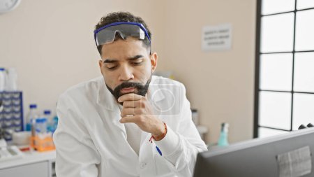 Photo for Thoughtful hispanic man with a beard in lab goggles contemplates in a laboratory environment. - Royalty Free Image