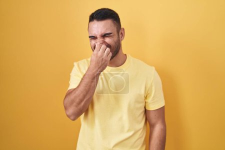 Téléchargez les photos : Jeune homme hispanique debout sur fond jaune sentant quelque chose de puant et dégoûtant, odeur intolérable, retenant souffle avec les doigts sur le nez. mauvaise odeur - en image libre de droit
