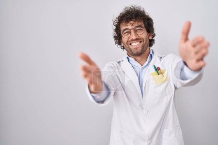 Téléchargez les photos : Jeune homme hispanique portant l'uniforme de médecin regardant la caméra souriant à bras ouverts pour étreindre. expression joyeuse embrassant le bonheur. - en image libre de droit
