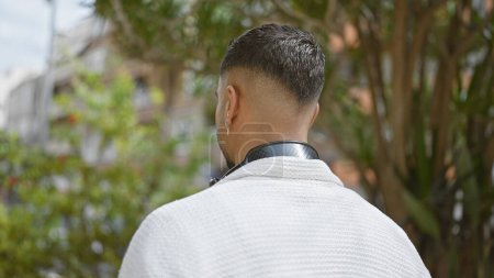 Téléchargez les photos : Vue arrière d'un jeune homme élégant avec une coiffure moderne et boucle d'oreille en plein air entourée de verdure. - en image libre de droit