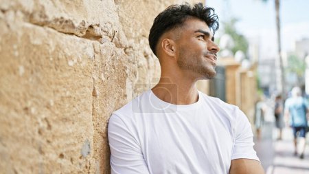 Hombre hispano guapo apoyado en una pared urbana iluminada por el sol al aire libre con la gente y la vida de la ciudad en el fondo