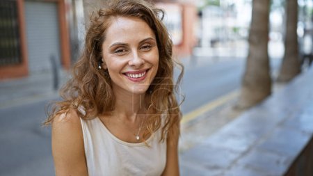 Photo for A smiling young woman with wireless earphones enjoys a sunny urban streetscape in the background. - Royalty Free Image