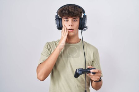 Téléchargez les photos : Adolescent hispanique jouant contrôleur de tenue de jeu vidéo peur et choqué, expression surprise et stupéfaite avec les mains sur le visage - en image libre de droit