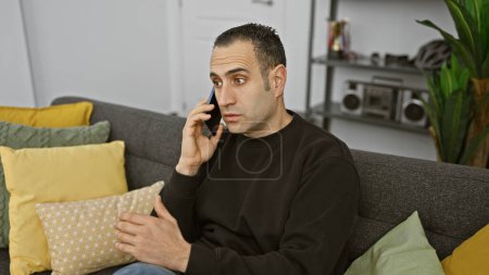 Téléchargez les photos : Bel homme hispanique parlant au téléphone dans un appartement moderne salon - en image libre de droit