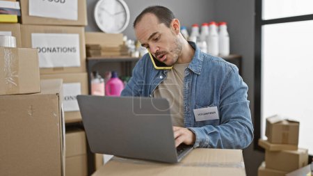 Hispanic man multitasking with phone and laptop in a warehouse surrounded by donation boxes.