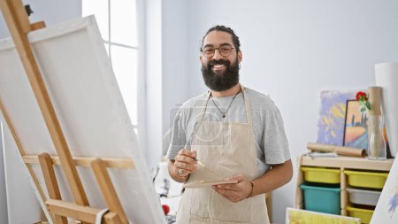 Photo for A cheerful young hispanic man with a beard and glasses, wearing an apron, paints in a bright art studio. - Royalty Free Image