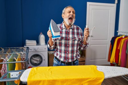 Wütender älterer Mann, wütend und wütend, schreit vor Wut, während er aggressiv in der Waschküche bügelt. unglücklicher Erwachsener, der starke Frustration ausstrahlt, inmitten der Hausarbeit schreit.
