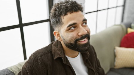 Téléchargez les photos : Jeune homme hispanique à la barbe souriante dans un salon moderne, évoquant des sentiments de confort, de bonheur et de style. - en image libre de droit