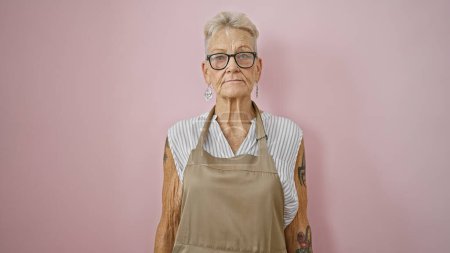 Foto de Mujer de pelo gris de mediana edad, un servidor de cara seria, está sola contra una vibrante pared rosa en su trabajo de camarera iluminada por el sol - Imagen libre de derechos