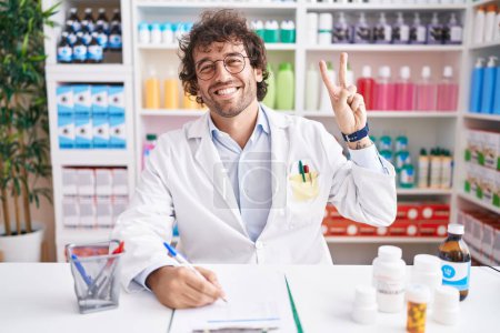 Téléchargez les photos : Jeune homme hispanique travaillant à la pharmacie pharmacie souriant en regardant la caméra montrant les doigts faisant signe de victoire. numéro deux. - en image libre de droit