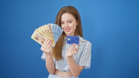 Téléchargez les photos : Jeune femme blonde souriante confiante tenant des billets de cent forints et carte de crédit sur fond bleu isolé - en image libre de droit