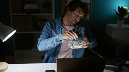 Téléchargez les photos : Homme hispanique mangeant de la salade dans un bureau sombre la nuit, travaillant tard dans les heures surlignées par la faible lumière de la lampe de bureau - en image libre de droit