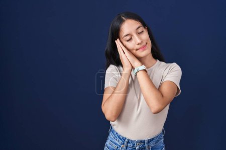 Foto de Young hispanic woman standing over blue background sleeping tired dreaming and posing with hands together while smiling with closed eyes. - Imagen libre de derechos
