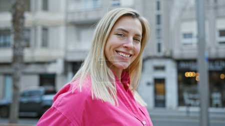 Téléchargez les photos : Jeune femme caucasienne souriante aux cheveux blonds portant une chemise rose, debout dans une rue urbaine. - en image libre de droit