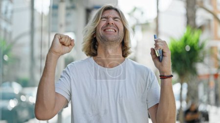 Photo for Ecstatic man with long blond hair celebrates outdoors in an urban setting, holding a smartphone. - Royalty Free Image