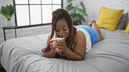 Foto de Relajada mujer afroamericana disfrutando de café de la mañana en la cama, rodeado de ambiente acogedor dormitorio - Imagen libre de derechos