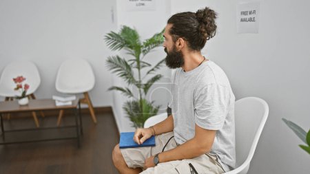 Téléchargez les photos : Bel homme hispanique avec une barbe assis dans une salle d'attente moderne tenant un carnet bleu - en image libre de droit