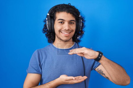 Photo for Hispanic man with curly hair listening to music using headphones gesturing with hands showing big and large size sign, measure symbol. smiling looking at the camera. measuring concept. - Royalty Free Image
