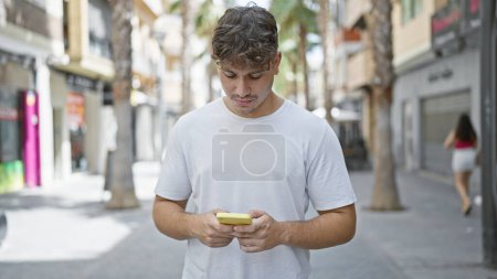 Photo for Young, handsome hispanic man, with a serious expression, engrossed in typing a message on his smartphone while standing under vibrant city sunlight, embodying a casual yet cool urban lifestyle. - Royalty Free Image