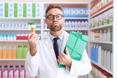 Téléchargez les photos : Homme d'âge moyen avec barbe travaillant à la pharmacie drugstore tenant des pilules en regardant la caméra souffler un baiser étant belle et sexy. expression d'amour. - en image libre de droit