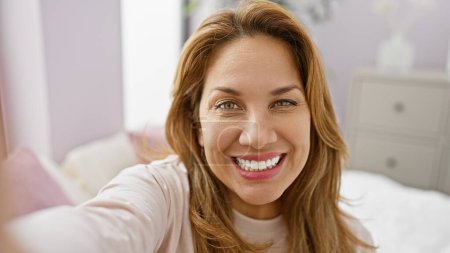 Téléchargez les photos : Une jeune femme hispanique joyeuse prend un autoportrait dans la chambre d'une maison confortable, incarnant la beauté et la positivité. - en image libre de droit