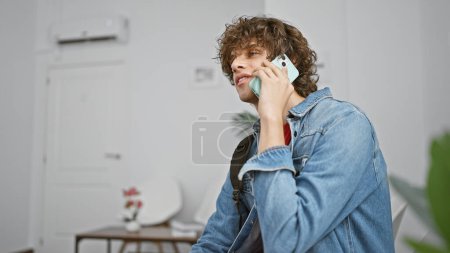 Photo for A young man with curly hair talks on a smartphone in a modern indoor space, giving off a casual and approachable vibe. - Royalty Free Image